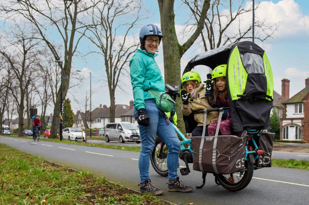 [photo of a smiling person with two kids on the back of their bike, on a cycle track separated from general road traffic]